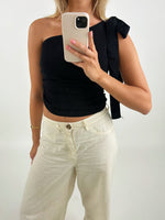 En person iført sort én-skulder top og hvide bukser tager et spejlbillede med en smartphone i hånden. Baggrunden er enkel og lys.