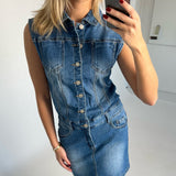 Kvinde i en ærmeløs denimkjole tager et spejlfoto af sig selv. Hun har rødmalede negle og et armbånd. Baggrunden er et lyst rum med simple vægge.