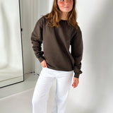 Kvinde iført brun sweatshirt og hvide bukser står afslappet i et lyst værelse, med en spejlvendt dør i baggrunden. Hun kigger let til siden.