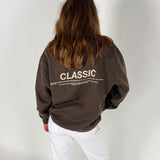 En person iført en brun sweatshirt med teksten "CLASSIC" og yderligere lille skrift. Personen står med ryggen til, klædt i hvide bukser, mod en hvid baggrund.