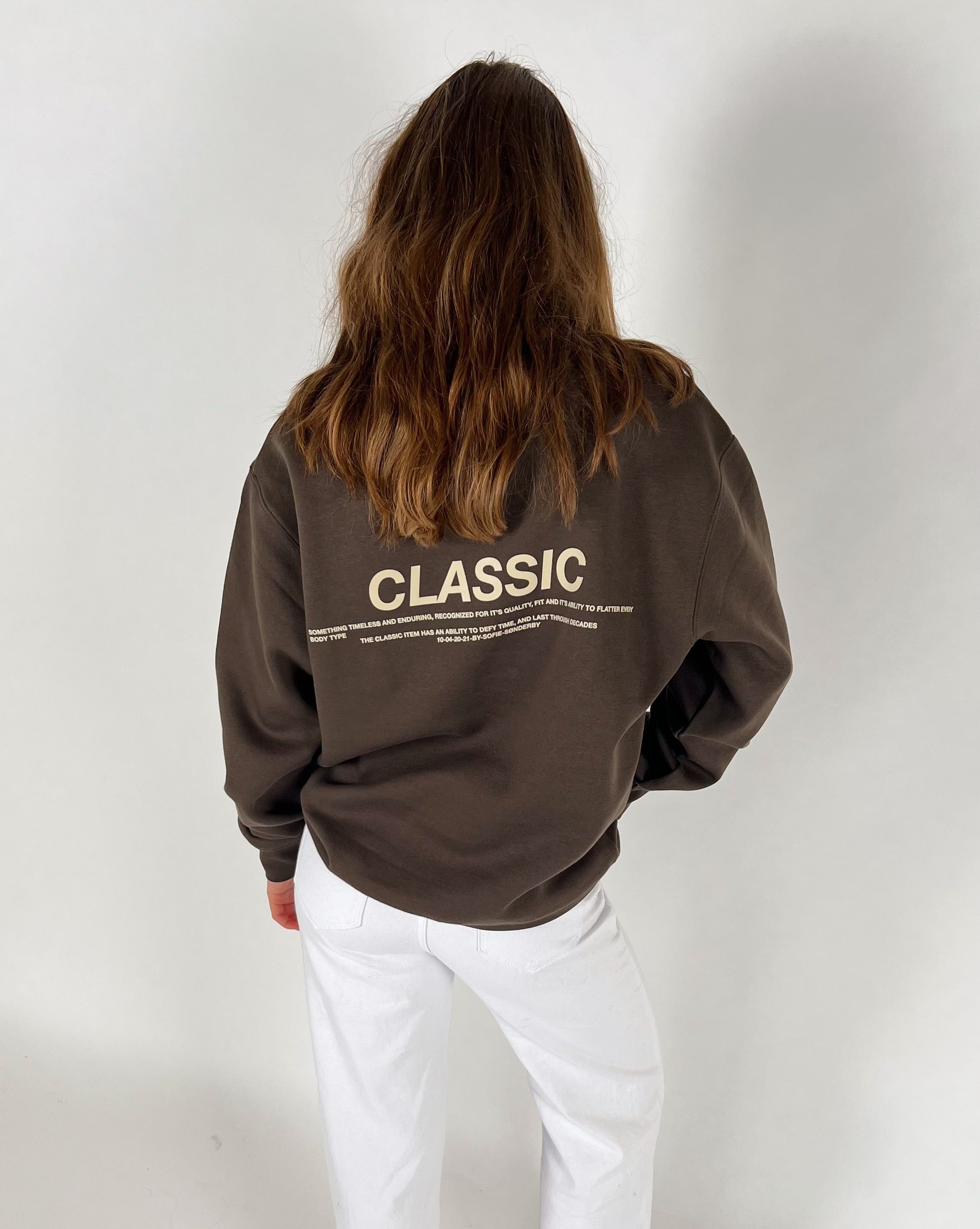 En person iført en brun sweatshirt med teksten "CLASSIC" og yderligere lille skrift. Personen står med ryggen til, klædt i hvide bukser, mod en hvid baggrund.