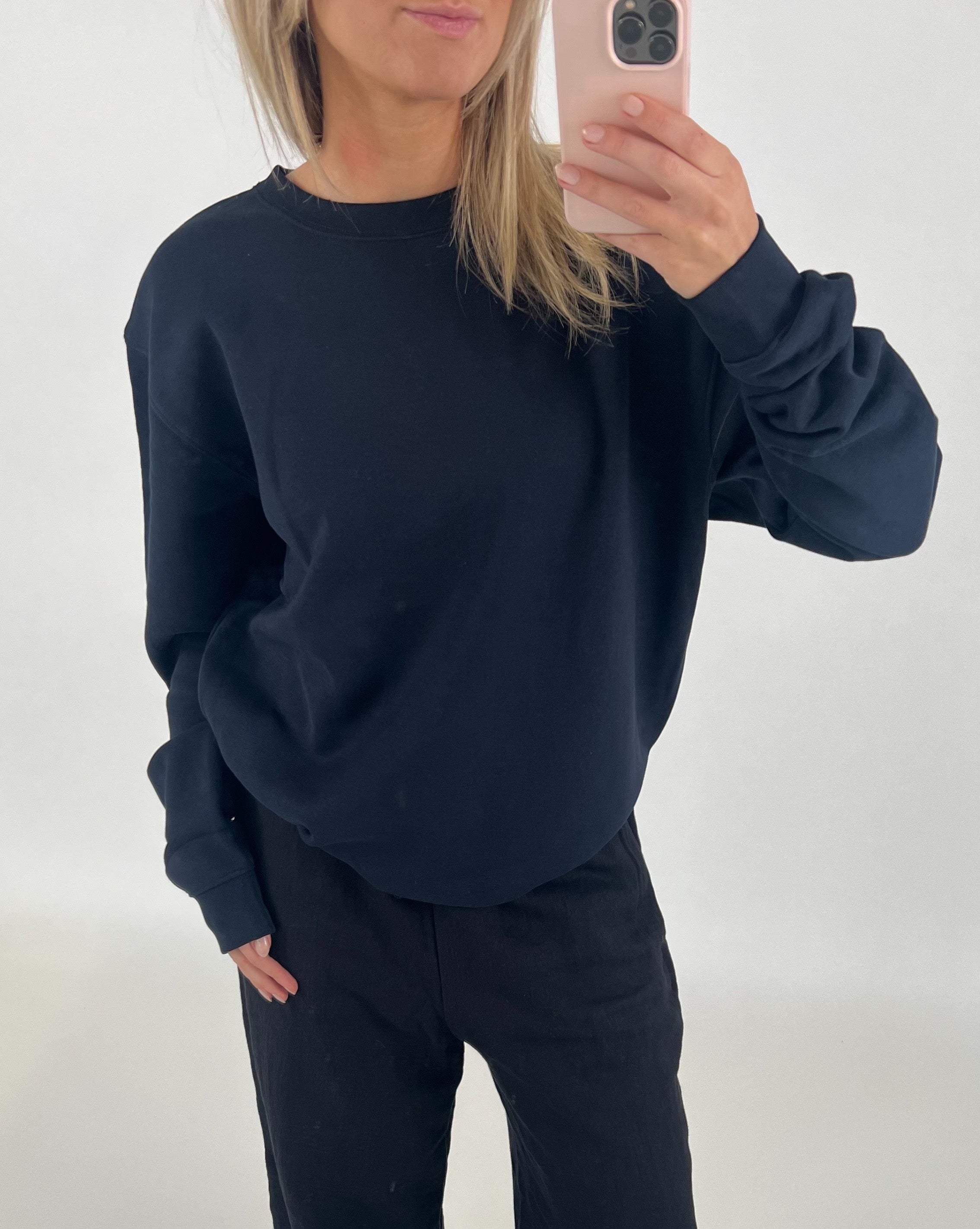 En person iført sort sweatshirt tager et spejlfoto med en smartphone i en lys, minimalistisk baggrund.