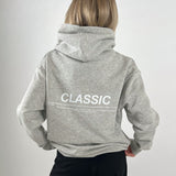 En person iført en grå hættetrøje med teksten "CLASSIC" står med ryggen til i en neutral, lys baggrund. Mere tekst: "SOMETHING TIMELESS AND ENDURING, RECOGNIZED FOR ITS QUALITY, FIT, AND ITS ABILITY TO…"