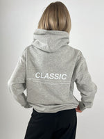 En person iført en grå hættetrøje med teksten "CLASSIC" står med ryggen til i en neutral, lys baggrund. Mere tekst: "SOMETHING TIMELESS AND ENDURING, RECOGNIZED FOR ITS QUALITY, FIT, AND ITS ABILITY TO…"