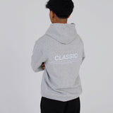 En person iført en grå hættetrøje med teksten "CLASSIC" står med krydsede arme i et hvidt rum.
