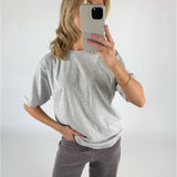 En person iført grå T-shirt og mørke bukser holder en smartphone foran ansigtet. Personen står i et lyst rum og ser ud til at tage et spejlbillede.