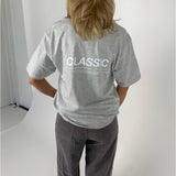 En person iført en grå T-shirt og mørke bukser står med ryggen til. På T-shirten står teksten "CLASSIC". Baggrunden er lys og enkel.