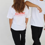 To personer iført hvide T-shirts og sorte bukser står mod en lys baggrund. Den ene T-shirt har tekst på ryggen: "CLASSIC" efterfulgt af yderligere ord i mindre skrift.