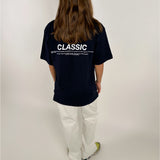 Person bærer en sort T-shirt med teksten "CLASSIC" i hvid, samt flere små skrifttyper nedenunder. Personen står med ryggen til iført hvide bukser og farverige sko i et neutralt rum.