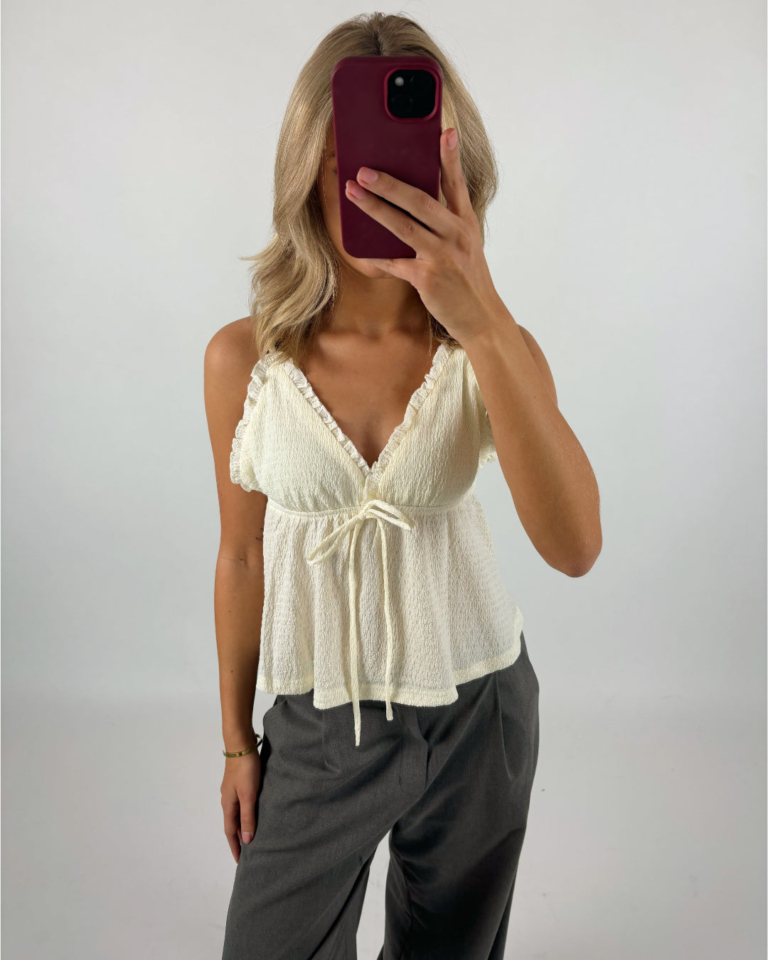 En person tager et spejlbillede med en smartphone med rød etui. Personen bærer en cremefarvet top med flæser og bindebånd samt grå bukser i et neutralt rum.