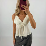 En person tager et spejlbillede med en smartphone med rød etui. Personen bærer en cremefarvet top med flæser og bindebånd samt grå bukser i et neutralt rum.