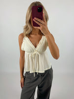 En person tager et spejlbillede med en smartphone med rød etui. Personen bærer en cremefarvet top med flæser og bindebånd samt grå bukser i et neutralt rum.