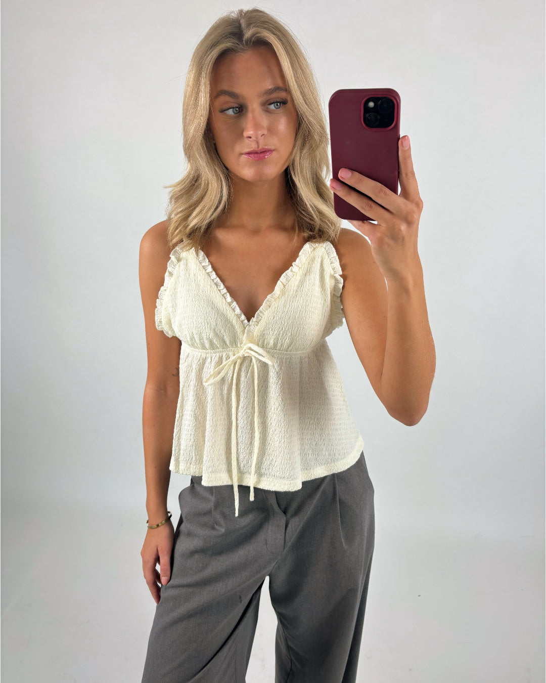 En person tager en selfie iført en lyseblå top og grå bukser. Personen står i et rum med en lys baggrund og holder en rød mobiltelefon.