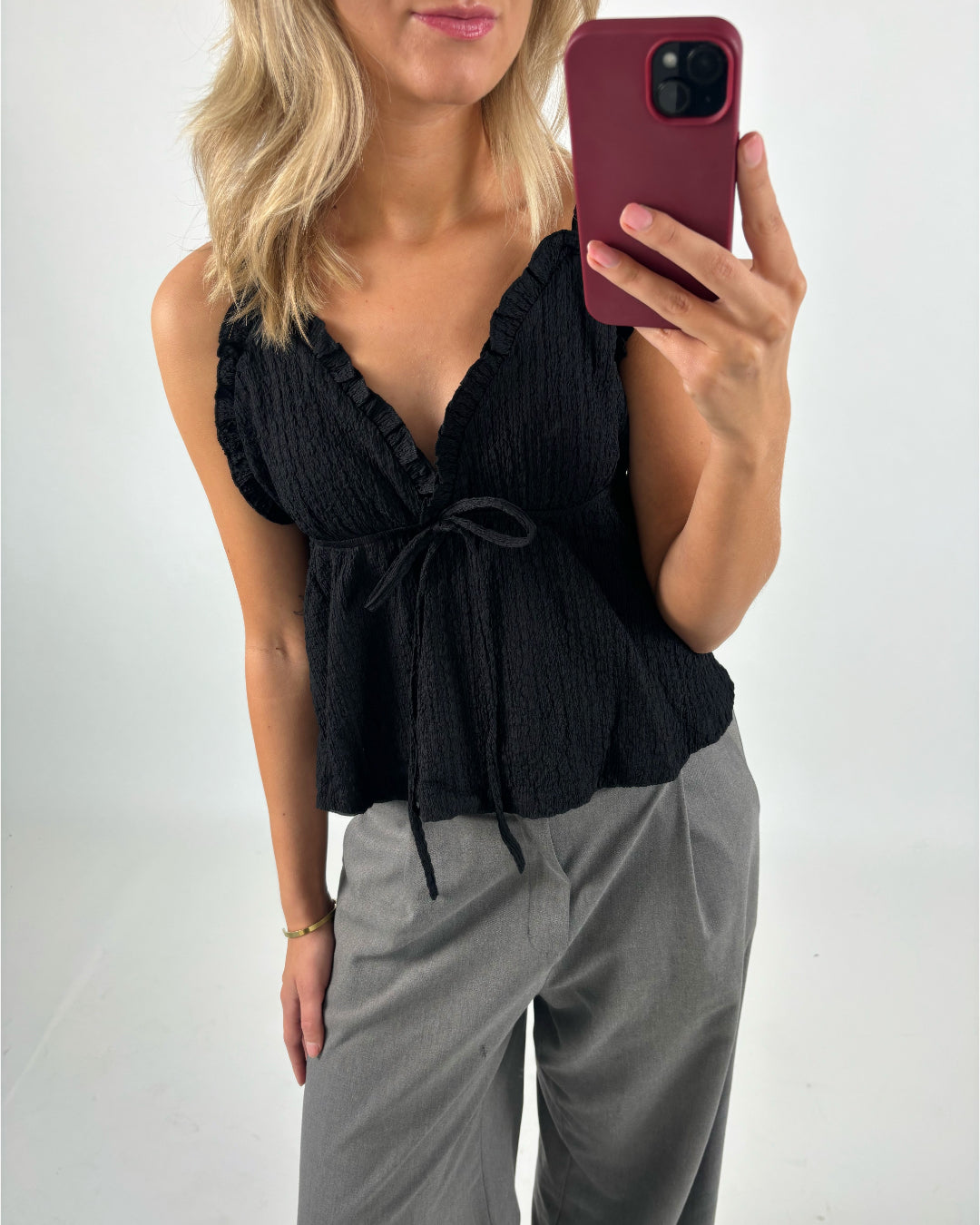 En person iført en sort bluse med flæsedetaljer og et bånd ses tage en selfie. Personen har blondt hår og bærer grå bukser. Baggrunden er neutral.