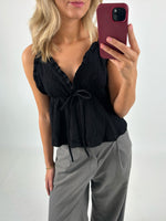 En person iført en sort bluse med flæsedetaljer og et bånd ses tage en selfie. Personen har blondt hår og bærer grå bukser. Baggrunden er neutral.