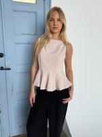 En kvinde står foran en lys blå dør iført en lys beige top og sorte bukser, med en neutral baggrund.