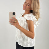 En kvinde i en hvid, mønstret bluse tager et spejlbillede med sin smartphone. Hun står i en minimalistisk indrettet lys baggrund. Hun bærer guldringe.