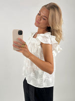 En kvinde i en hvid, mønstret bluse tager et spejlbillede med sin smartphone. Hun står i en minimalistisk indrettet lys baggrund. Hun bærer guldringe.