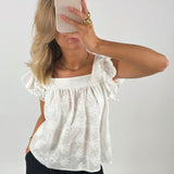 En person iført hvid, mønstret bluse tager et spejlselfie med en smartphone. Personen har let bølget, blond hår og guldtilbehør. Baggrunden er en ensfarvet væg.