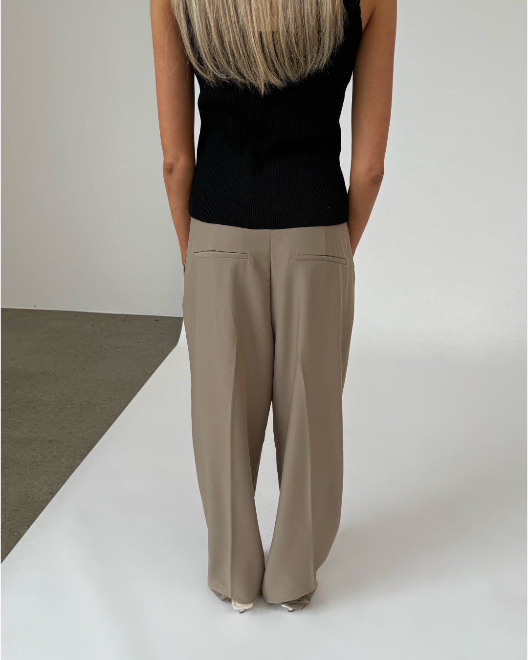 En person står med ryggen til, iført en sort top og beige bukser. Vedkommende har langt blondt hår, og baggrunden er en lys, minimalistisk indendørs setting.