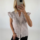 Kvinde iført blomstret bluse tager selfie med mobiltelefon i et lyst rum. Hun har sort tøj på og bærer et guldarmbånd.