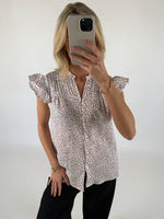 Kvinde iført blomstret bluse tager selfie med mobiltelefon i et lyst rum. Hun har sort tøj på og bærer et guldarmbånd.