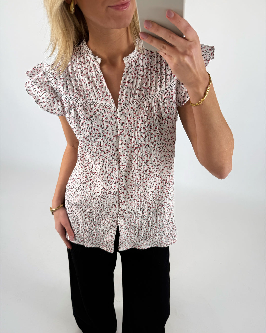 En person iført en blomstret skjorte tager et spejlbillede med en smartphone. Personen bærer også sorte bukser og et armbånd, og står i et lyst rum.