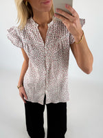 En person iført en blomstret skjorte tager et spejlbillede med en smartphone. Personen bærer også sorte bukser og et armbånd, og står i et lyst rum.