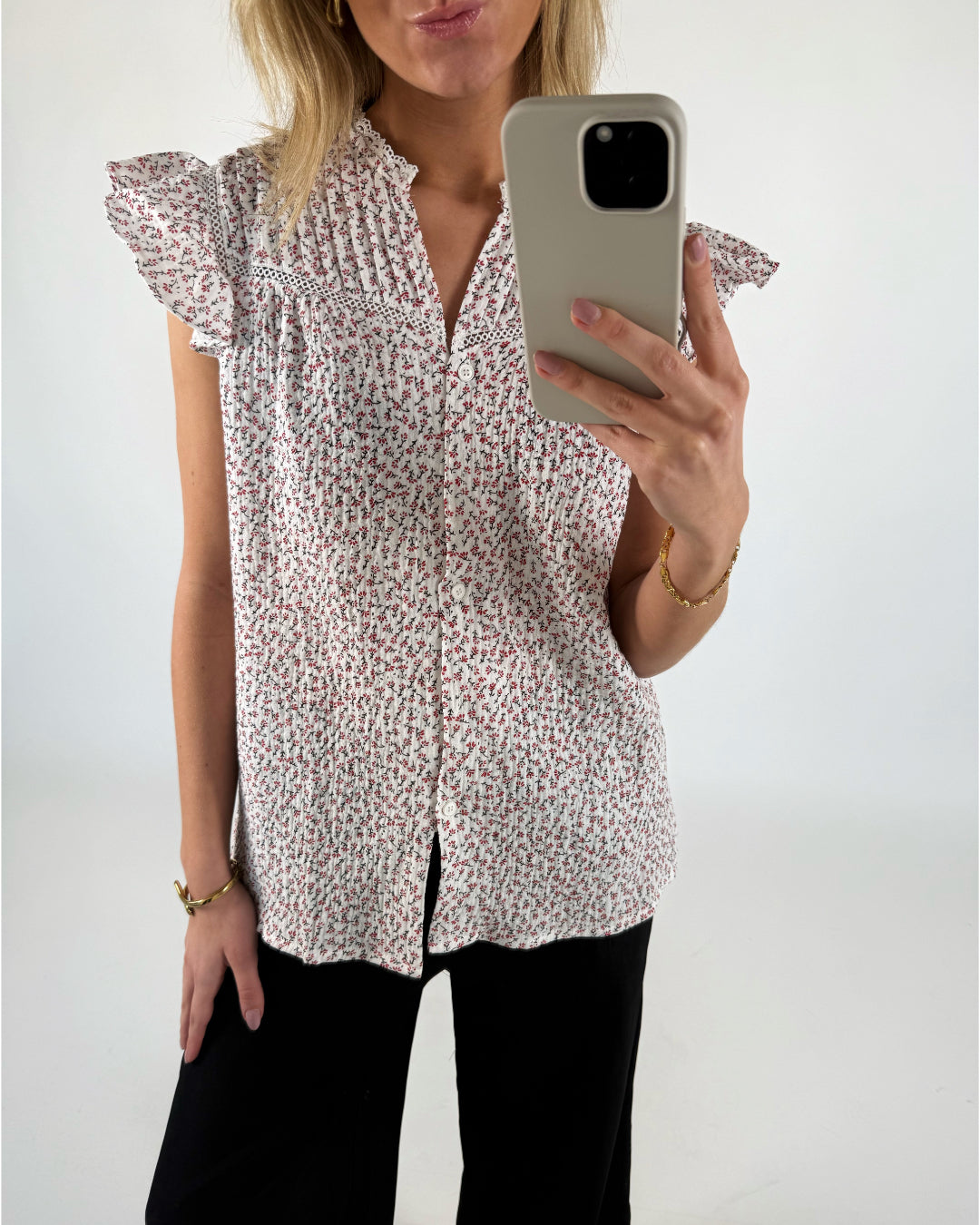 Kvinde iført blomstret skjorte med korte ærmer tager et spejlbillede med mobiltelefonen. Hun står i et hvidt rum og bærer sort bukser og et guldarmbånd.