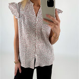 Kvinde iført blomstret skjorte med korte ærmer tager et spejlbillede med mobiltelefonen. Hun står i et hvidt rum og bærer sort bukser og et guldarmbånd.