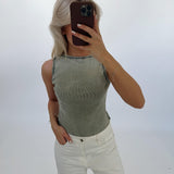 En person tager et selfie iført en lysegrøn top og hvide bukser. Personen holder en mobiltelefon foran ansigtet i et neutralt belyst rum.