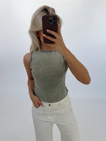 En person tager et selfie iført en lysegrøn top og hvide bukser. Personen holder en mobiltelefon foran ansigtet i et neutralt belyst rum.