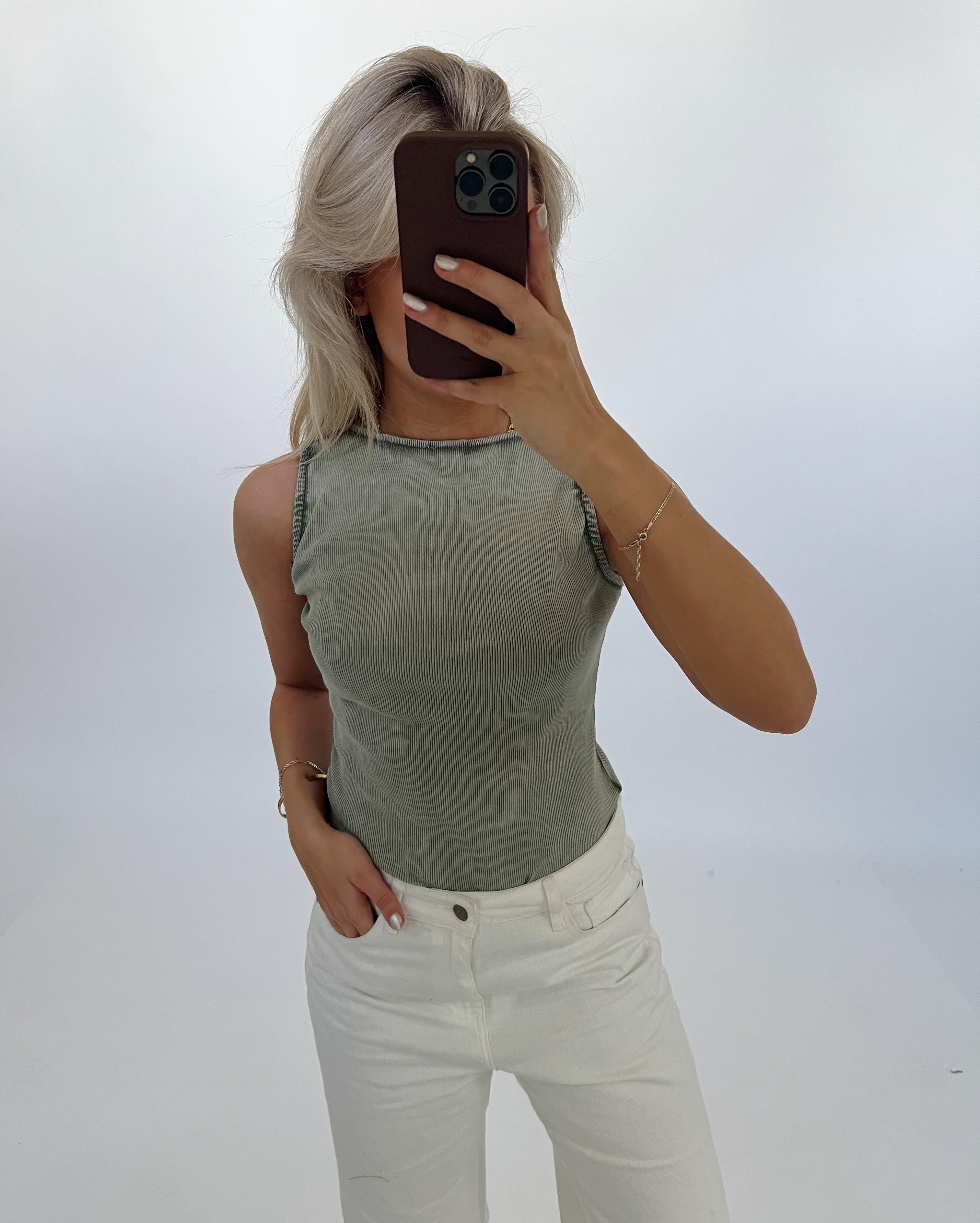 En person tager et selfie iført en lysegrøn top og hvide bukser. Personen holder en mobiltelefon foran ansigtet i et neutralt belyst rum.