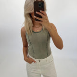 Person tager et spejlfoto med mobiltelefon, iført en grøn top og hvide bukser. Baggrunden er en lys, tom væg.