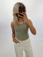 Person tager et spejlfoto med mobiltelefon, iført en grøn top og hvide bukser. Baggrunden er en lys, tom væg.