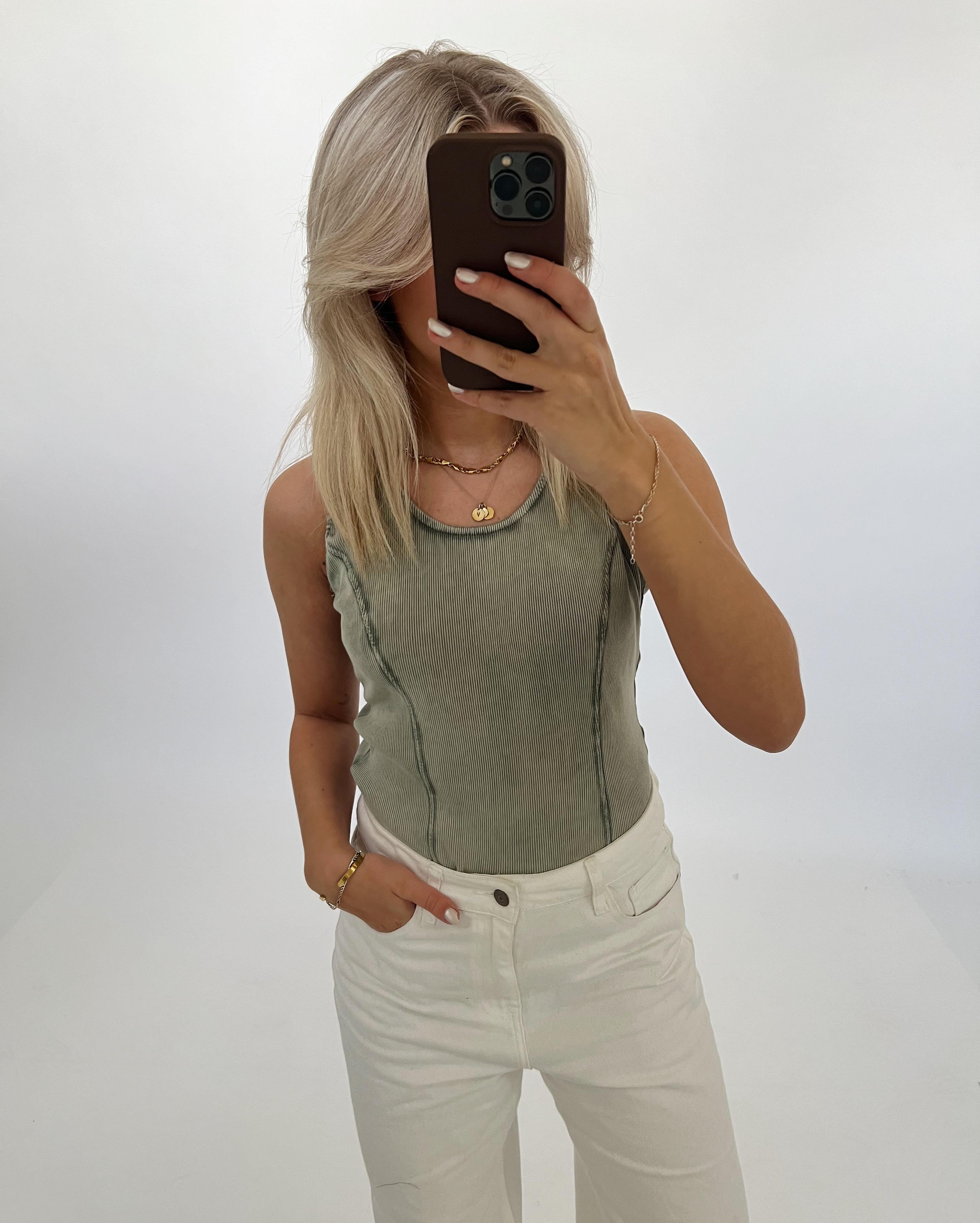 Person tager et spejlfoto med mobiltelefon, iført en grøn top og hvide bukser. Baggrunden er en lys, tom væg.