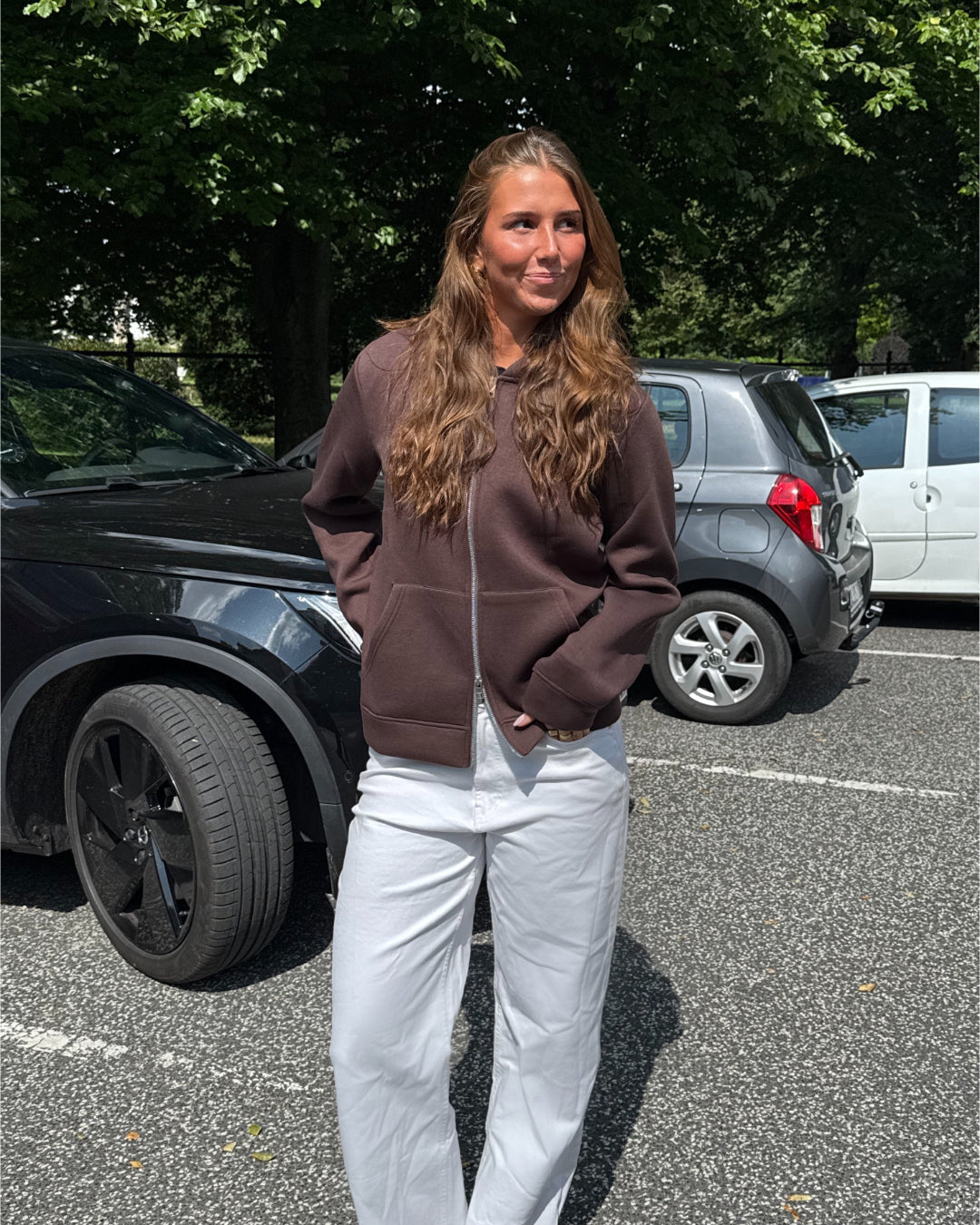 Modelfoto af pige, som bærer EMMA hoodie, brun og NANNA jeans, hvid - alt fra BySofieSønderby. Baggrund er p-plads og træer