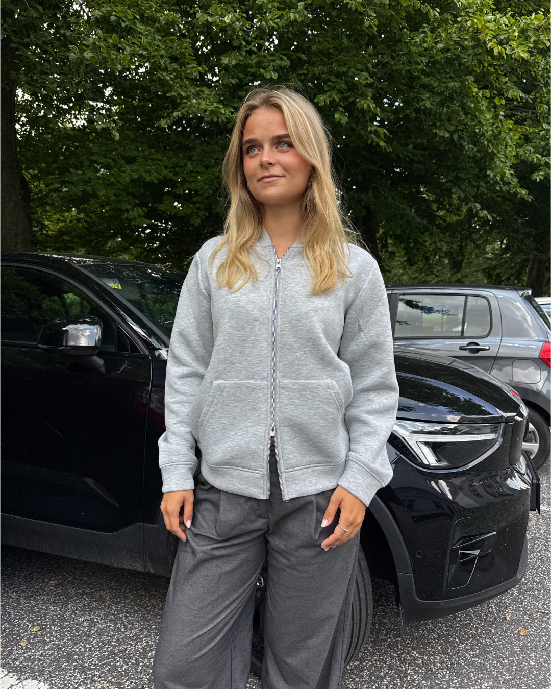 Modelfoto af pige, som bærer EMMA hoodie, lysegrå og MILAN bukser, grå - alt fra BySofieSønderby. Baggrund er p-plads og træer