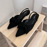 Sort slingback-sko med sløjfer placeret stilfuldt på en lys træhylde i et minimalistisk rum. Skoene har teksten "soft mix" på sålerne.