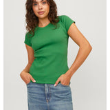 En person står iført en grøn t-shirt og blå jeans, med hænderne i lommerne, mod en lys baggrund. Persoen har krøllet hår og ser direkte fremad.
