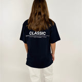 Person i sort T-shirt står med ryggen til, iført hvide bukser. T-shirten har teksten: "CLASSIC" efterfulgt af mindre tekst. Baggrunden er lys og enkel.