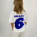 En person ses bagfra i en hvid T-shirt med blå tekst "GALAXY 6" i et lyst rum. Personen står stille med hænderne ved siden.