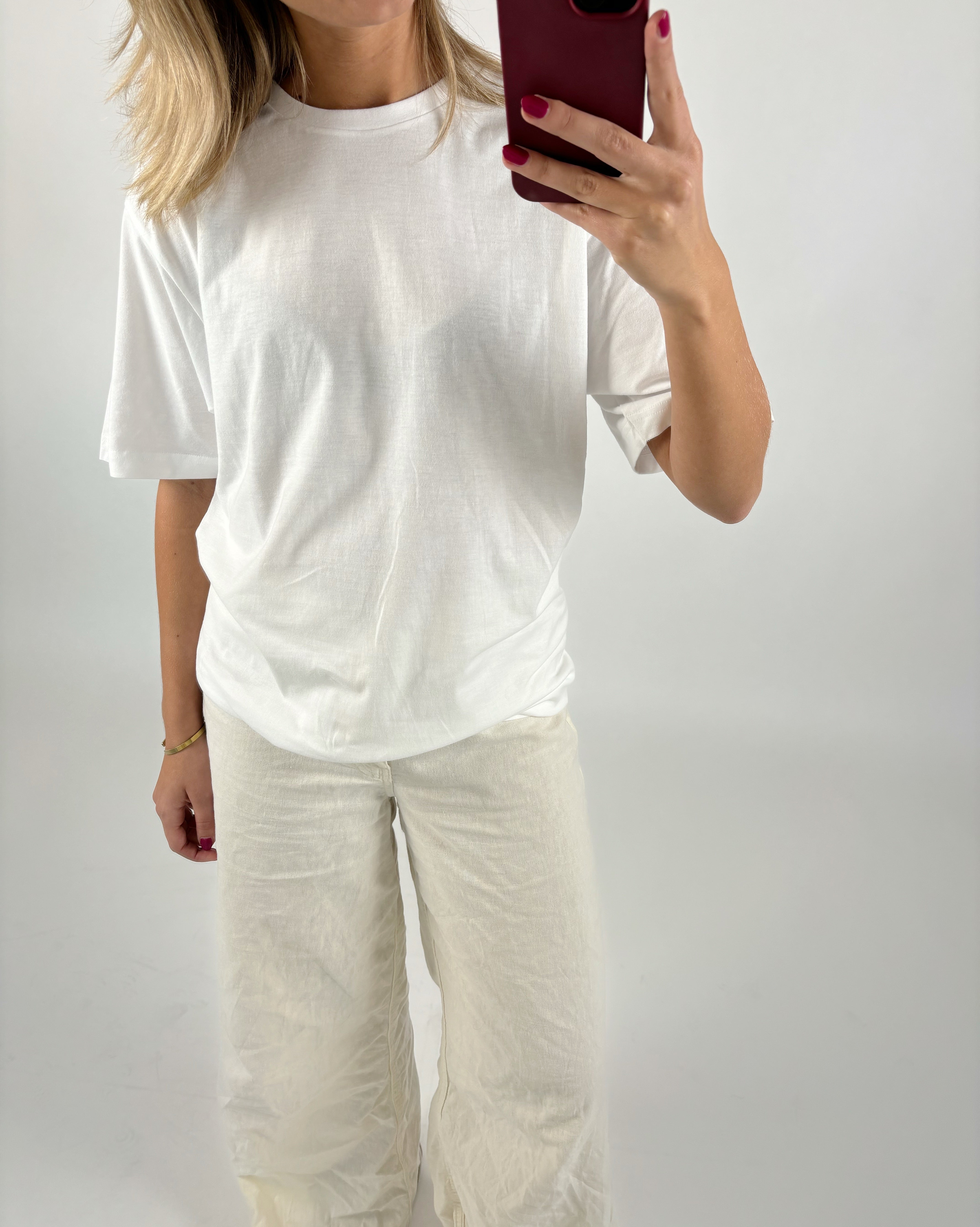En person iført en hvid T-shirt og hvide bukser tager et spejlbillede med en mobiltelefon i et simpelt, neutralt rum.