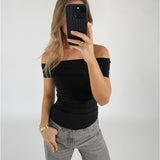 Person tager et spejlbillede iført en sort off-shoulder-top og grå bukser, i et enkelt rum. Personen holder en sort telefon op til ansigtet.