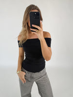 Person tager et spejlbillede iført en sort off-shoulder-top og grå bukser, i et enkelt rum. Personen holder en sort telefon op til ansigtet.