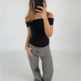 En person tager et spejlbillede, iført en sort, skulderfri top og grå jeans, mens de står i et minimalistisk, lyst rum med en neutral væg.