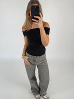 En person tager et spejlbillede, iført en sort, skulderfri top og grå jeans, mens de står i et minimalistisk, lyst rum med en neutral væg.