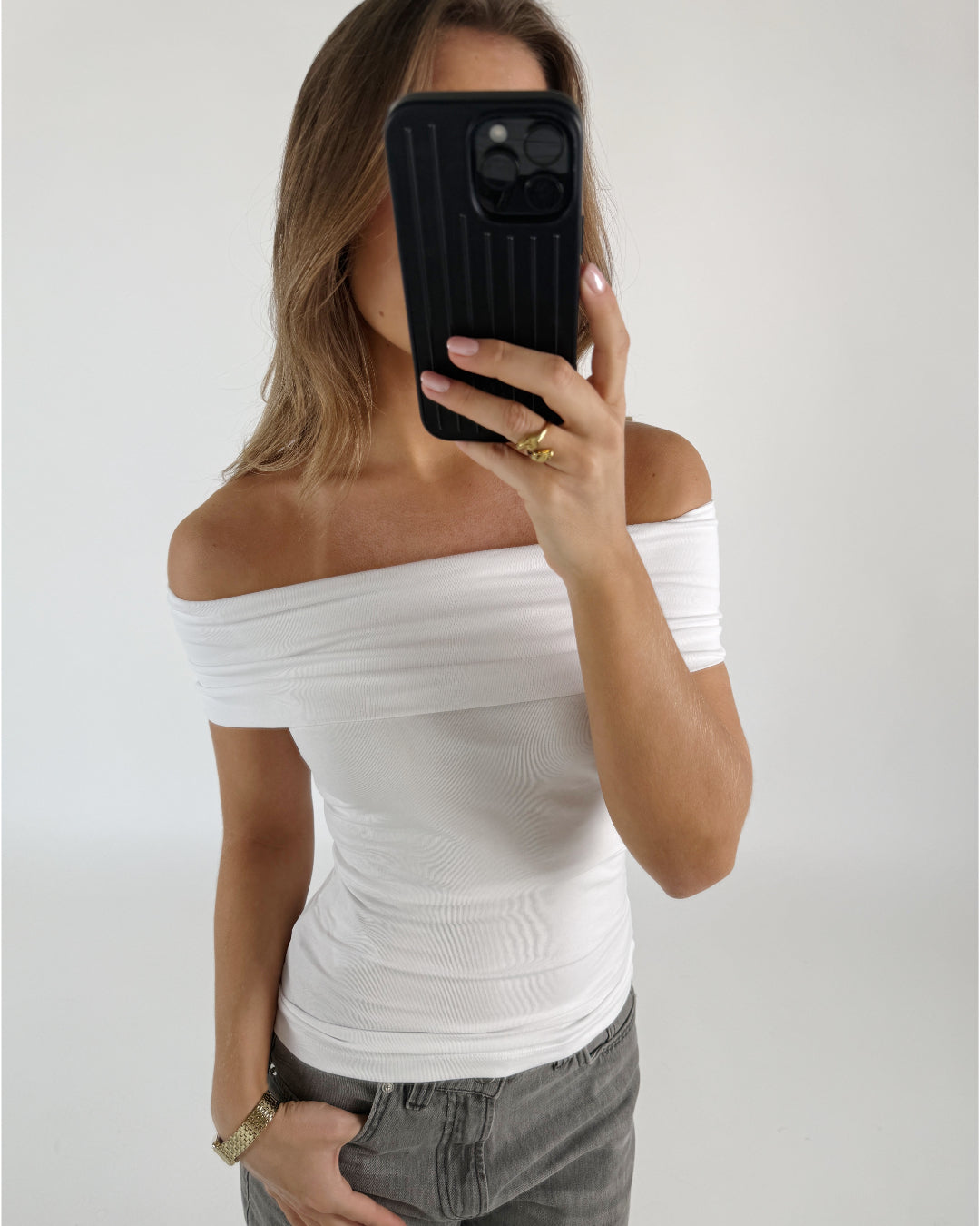 En person med en hvid off-shoulder top holder en sort smartphone foran ansigtet. Personen står i et lyst rum, iført grå jeans og en guld ring.