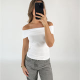 Person iført en hvid off-shoulder top og grå bukser tager et spejlfoto med en sort telefon. Baggrunden er en simpel, hvid væg.