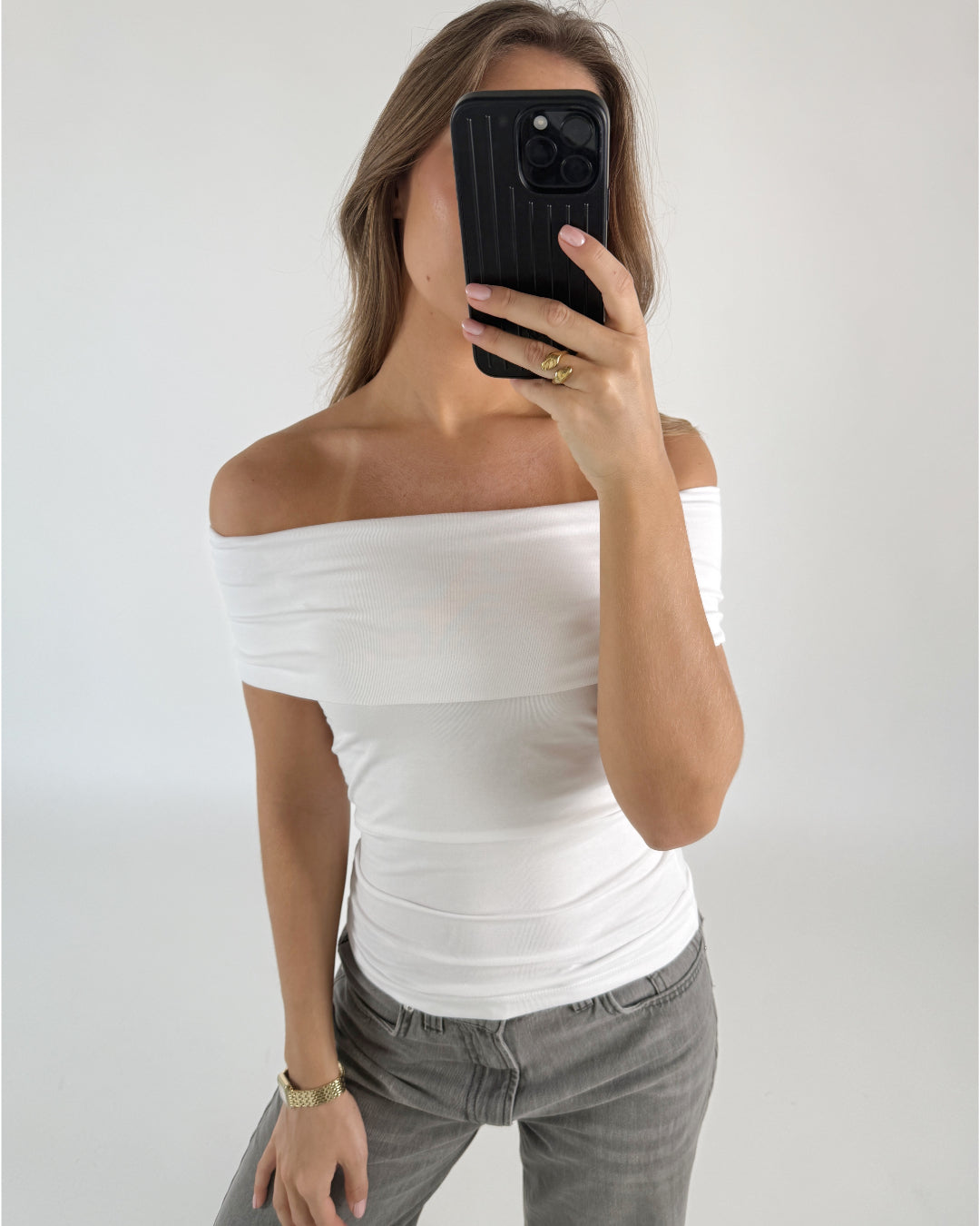 Person i hvid top tager et billede med en sort telefon, iført grå jeans og smykker, står i et minimalistisk rum.